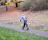 A person in a blue hoodie and dark denim jeans walking along a park path, they are hunched over at the waist.